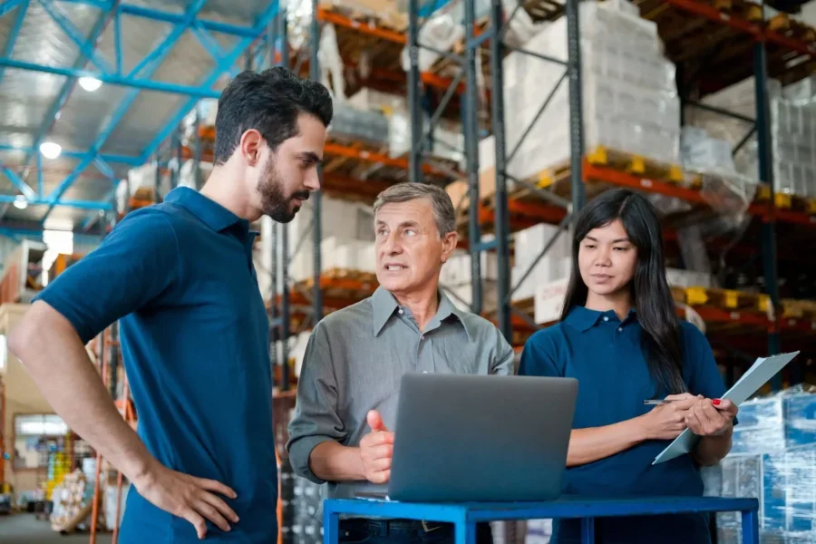 Male senior manager wearing grey shirt standing in warehouse, using laptop and talking with employees.