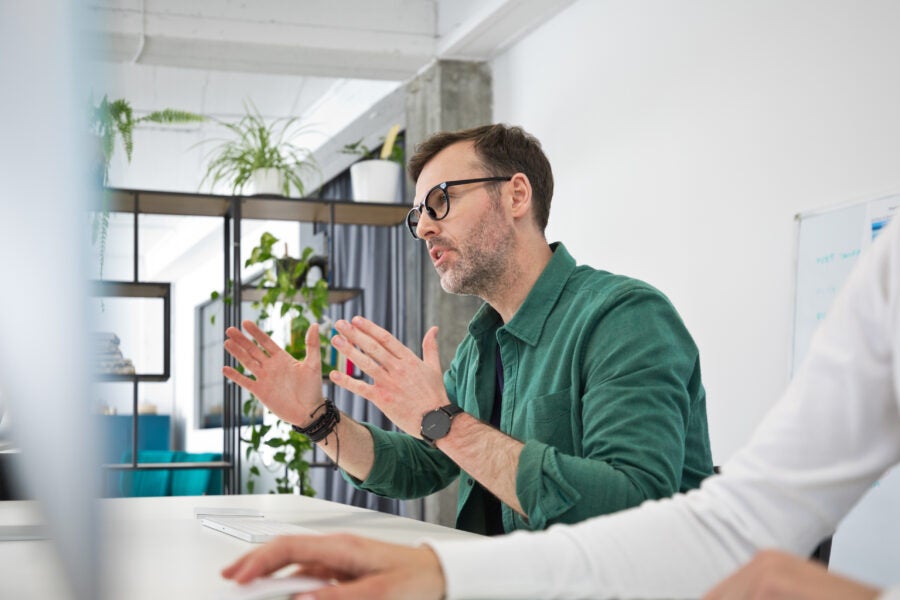 Mature man wearing green shirt and eyeglasses sitting at the desk in the office, using computer, talking and gesturing. Businessman during video conference.