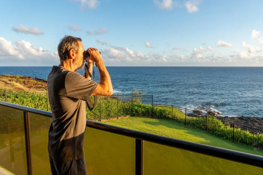 Mature Caucasian man using binoculars standing on a balcony over the ocean and green grass and iron fence, Makahuena Point, Kauai, Hawaii
