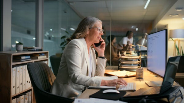 Computer, night and phone call with mature woman in office for budget or finance update. Accounting, bookkeeping and communication with business employee typing in workplace for financial consulting