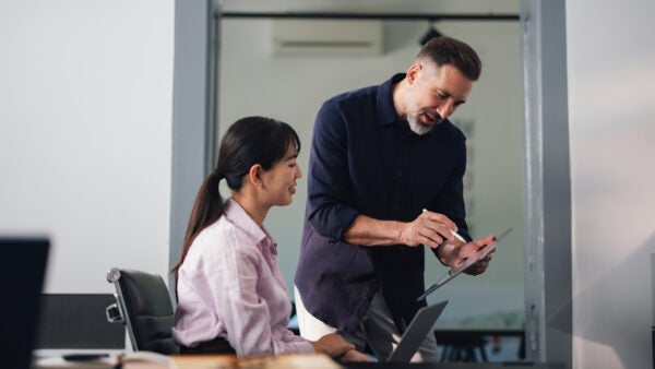 A seasoned man explains a tablet to an attentive woman in a contemporary office, capturing teamwork, mentorship, and professional collaboration in a coworking environment.