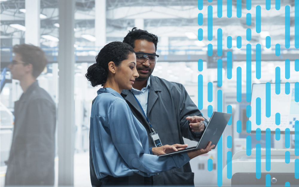 A manager and an engineer review information on a laptop together on a manufacturing facility floor.