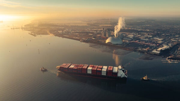Aerial view of a container ship moving through an estuary at sunrise, with an industrial port and facility on the far shore.