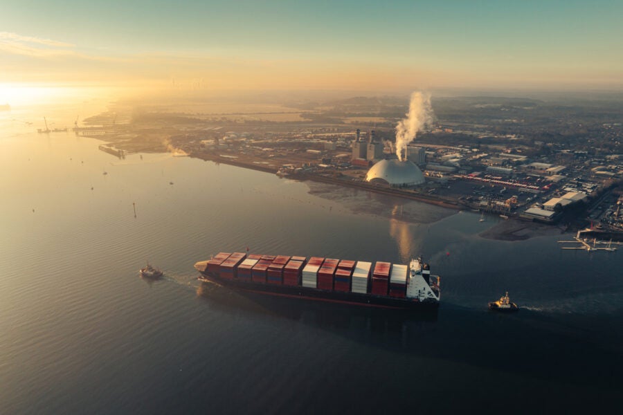 Aerial view of a container ship moving through an estuary at sunrise, with an industrial port and facility on the far shore.